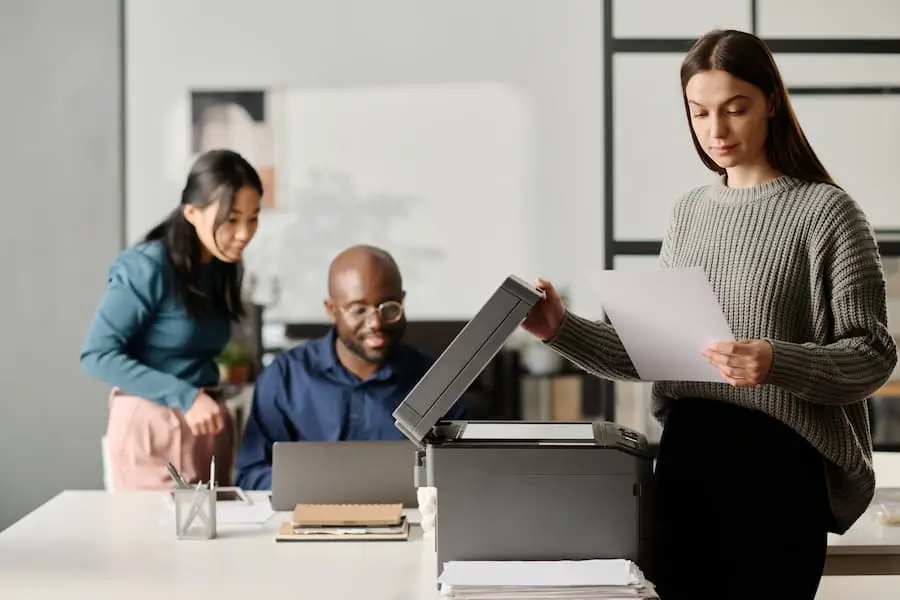 A woman in a gray sweater using a copier while two colleagues, a man and a woman, work and converse in the background at an office.